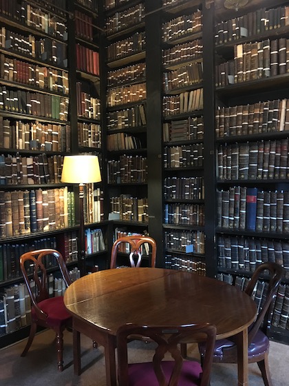 Inside the Portico Library Manchester. Floor to ceiling bookshelves, low lighting. A round table with 4 chairs in the foreground and a lamp in the corner.